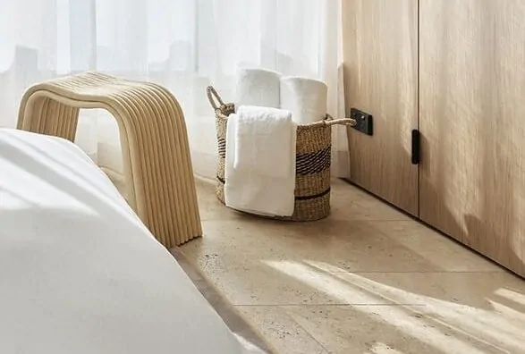 A warm, minimalist bedroom with travertine flooring, a woven stool, and soft natural light.