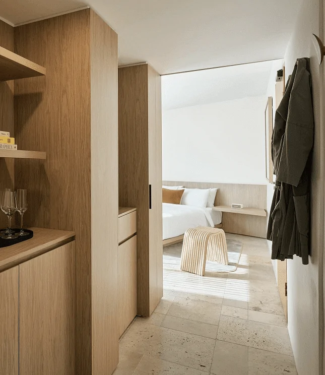 A warm, modern bedroom entrance with travertine flooring, wooden cabinetry, and a bright, minimalist sleeping area in view.