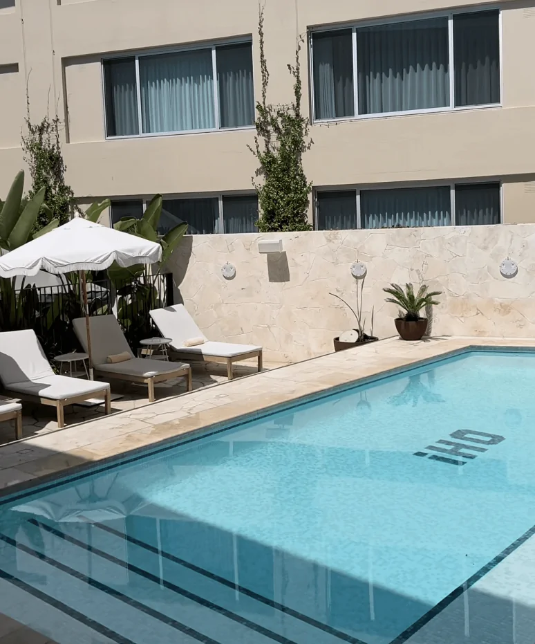 Poolside setting with travertine decking, lounge chairs, white umbrellas, and a modern hotel facade in the background.